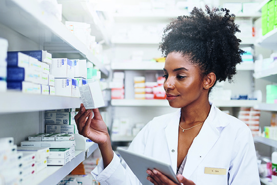 Ärztin in Praxisapotheke mit Medikamentenpackung in der Hand gleicht Bestände am Tablet ab.